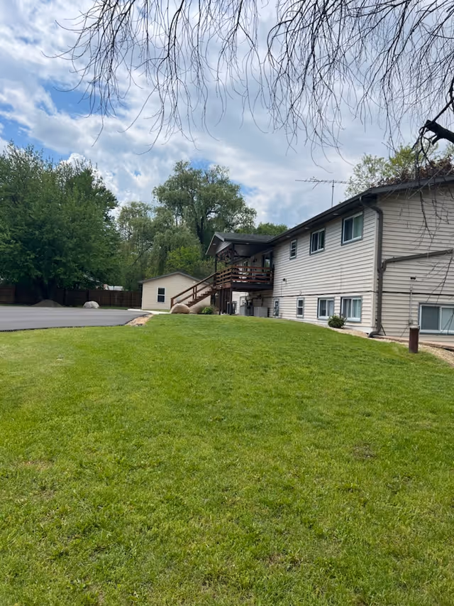 Exterior view of a two-story beige building with multiple windows, a wooden staircase and balcony, surrounded by green grass and trees under a partly cloudy sky.