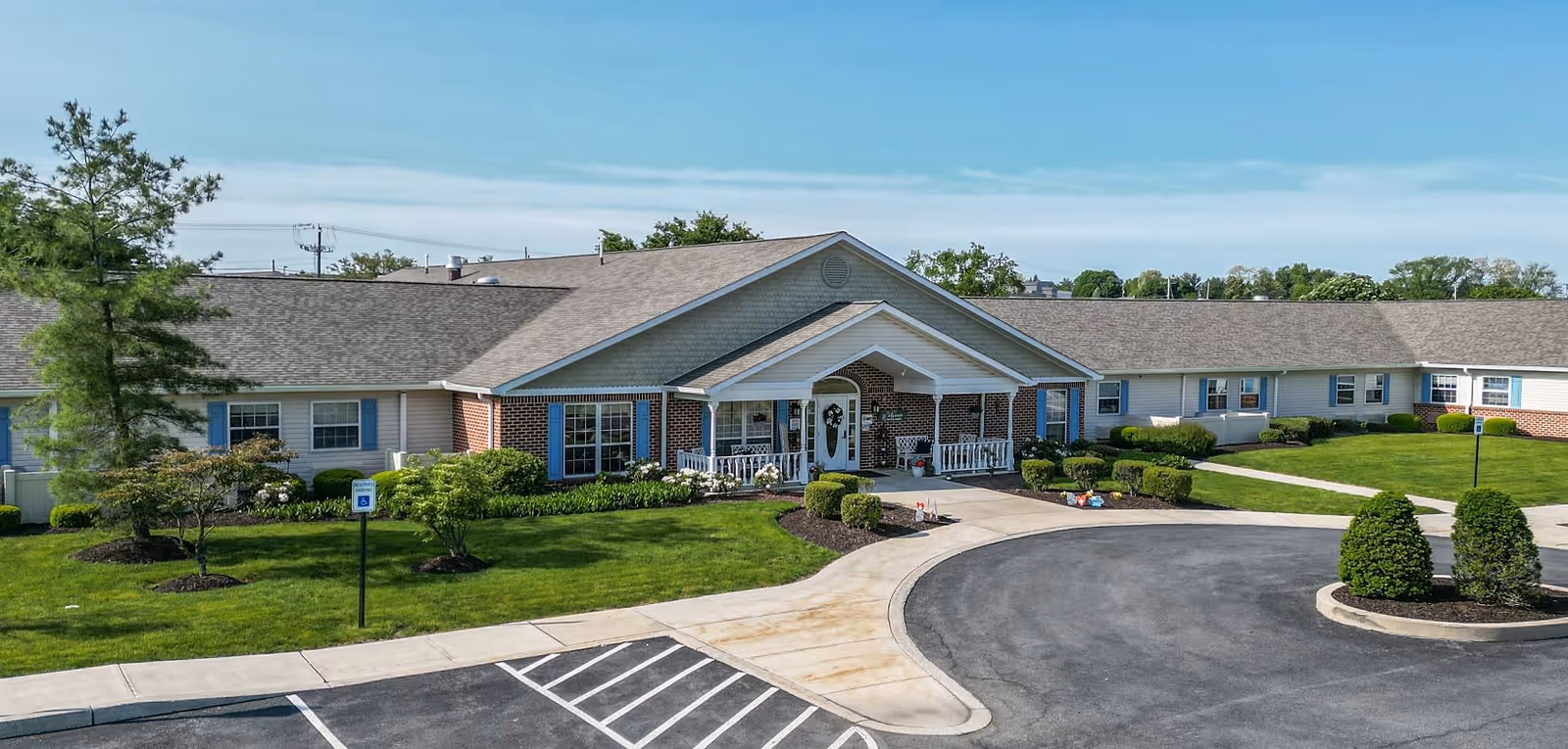 Exterior view of Arden Courts - ProMedica Memory Care Community (Susquehanna), a single-story brick and siding building with a covered entrance, surrounded by well-maintained green lawns, shrubs, and trees under a clear blue sky. There is a paved driveway and parking area in front.
