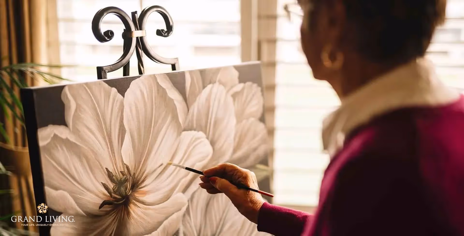 An elderly person painting a large white flower on a canvas set on an easel, with sunlight coming through window blinds in the background.