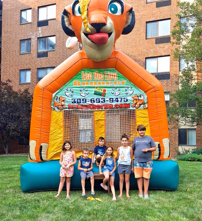 A group of children stand and sit in front of a large orange "Leo the Lion" inflatable bouncer on the lawn outside a brick building.