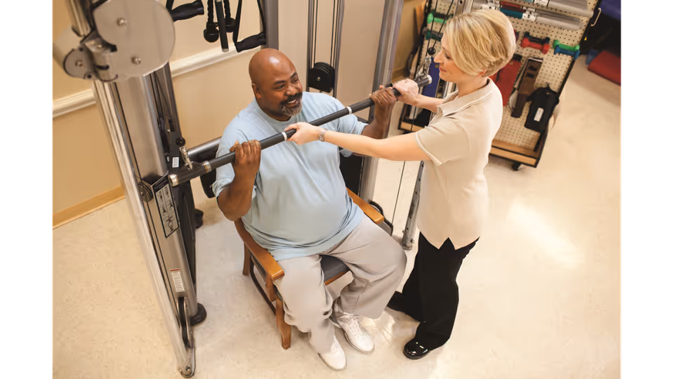 A man seated on a chair is using a weightlifting machine with assistance from a woman standing beside him, helping him hold the bar. The setting appears to be a rehabilitation or physical therapy room with exercise equipment and accessories visible in the background.