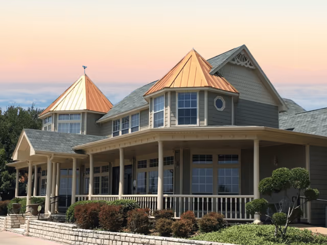 Front exterior of a two-story senior living building with a wraparound porch, copper-roofed turrets, and landscaped shrubs.