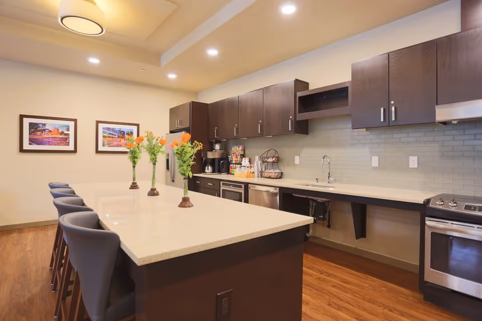 Modern kitchen area in a senior living facility with a large island countertop surrounded by chairs, three vases with flowers on the island, dark wood cabinets, stainless steel appliances including a refrigerator, dishwasher, and stove, and two framed pictures on the wall.