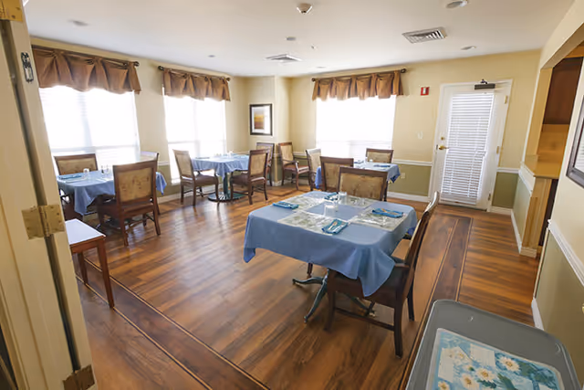 A bright dining room with several tables covered in blue tablecloths, each set with plates, glasses, and napkins. The room has wooden floors, multiple windows with brown valances, and wooden chairs around the tables. There is a door with a window blind and a small side table with a floral tray in the foreground.