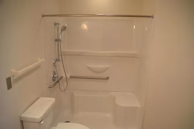 Bathroom with a white bathtub/shower unit featuring a handheld showerhead, horizontal grab bar and built-in seat, with a toilet to the left.
