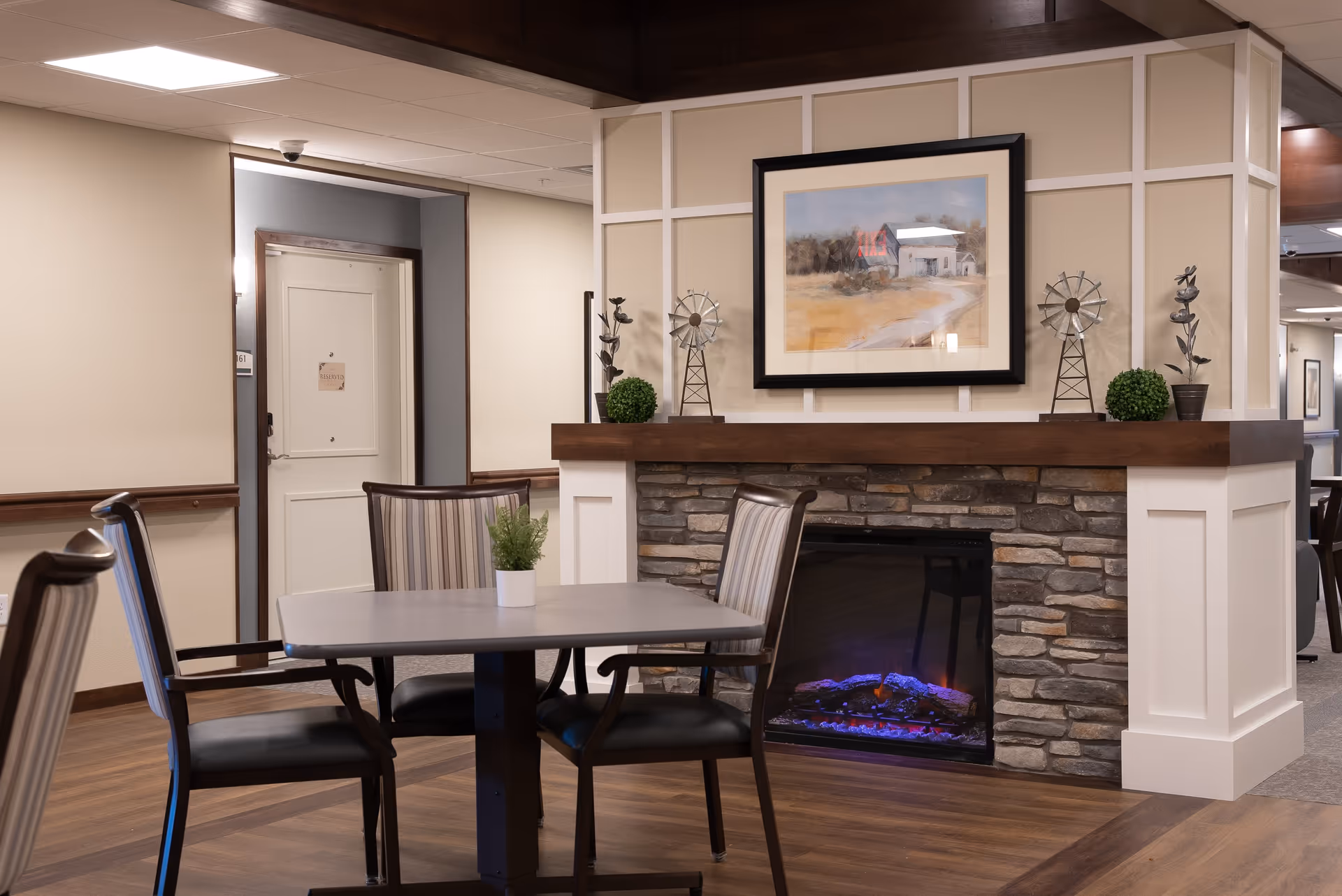 A cozy senior living common area with a table and chairs in front of a stone fireplace topped with framed artwork and decorative accents.