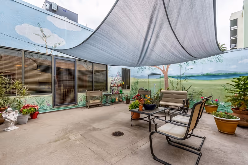 Outdoor patio area with a large gray shade sail overhead, surrounded by walls painted with a mural of trees, grass, and a lake. The patio has several potted plants, a small table with chairs, and a bench. There is a glass door and windows with blinds on one side.