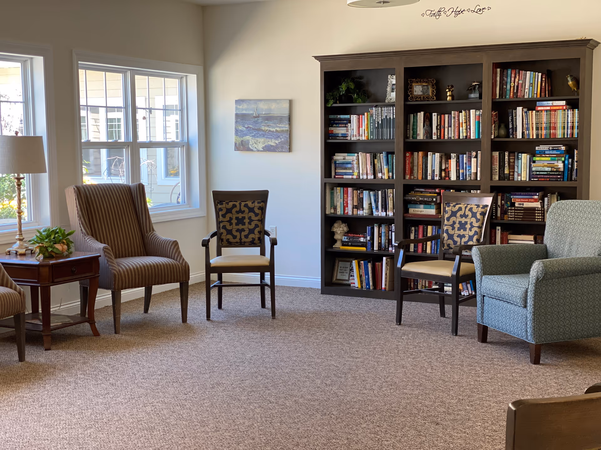 A cozy living room area with a large dark wood bookshelf filled with books, several upholstered chairs including a striped armchair, two patterned wooden chairs, and a blue armchair. There is a wooden side table with a lamp and a small plant next to a window letting in natural light. A small painting hangs on the wall above an electrical outlet.