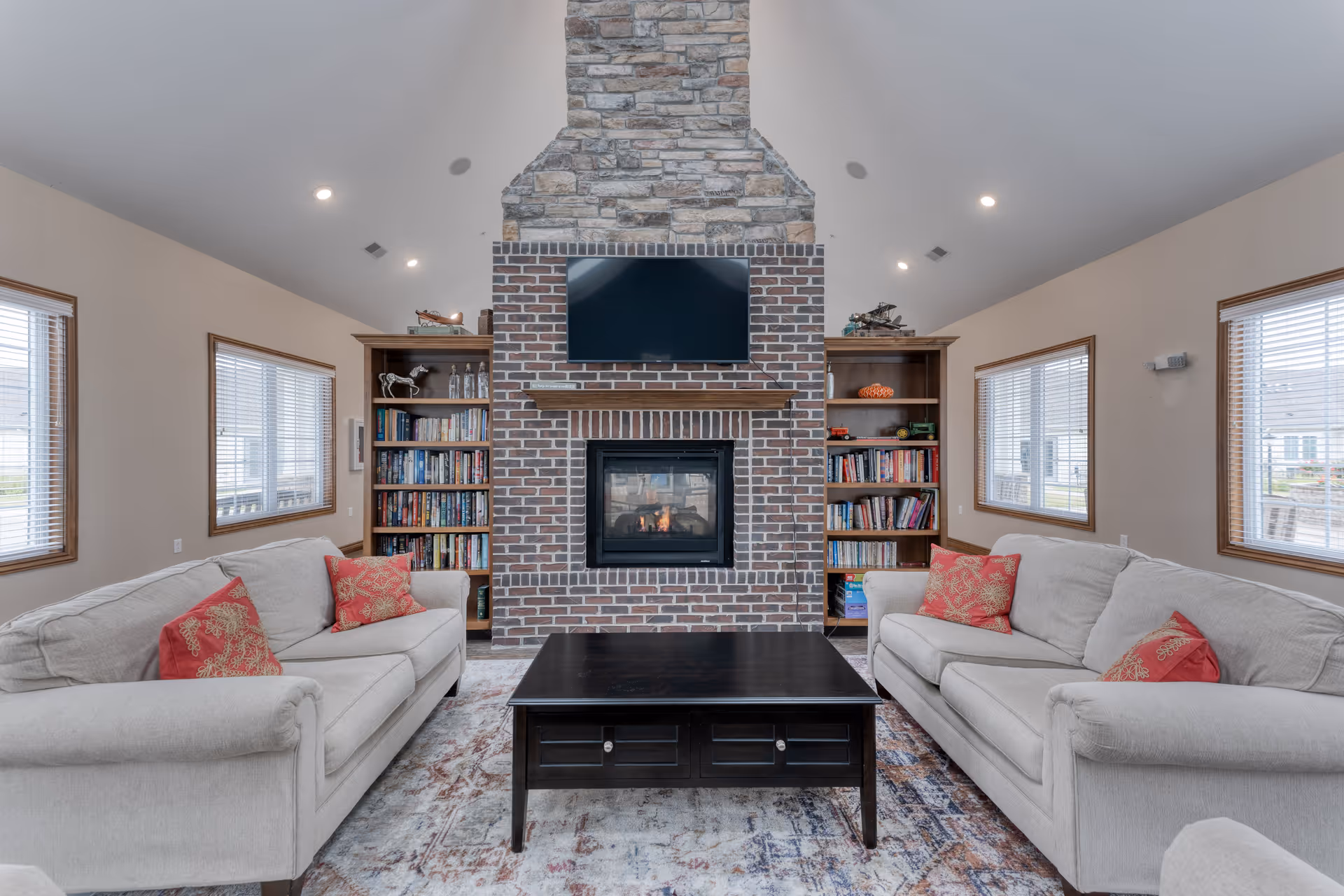 A cozy living room with two beige sofas facing each other, each adorned with red patterned throw pillows. Between the sofas is a dark wooden coffee table. The room features a large brick fireplace with a stone chimney, above which is a mounted flat-screen TV. On either side of the fireplace are wooden bookshelves filled with books and decorative items. The room has multiple windows with wooden frames allowing natural light to enter.