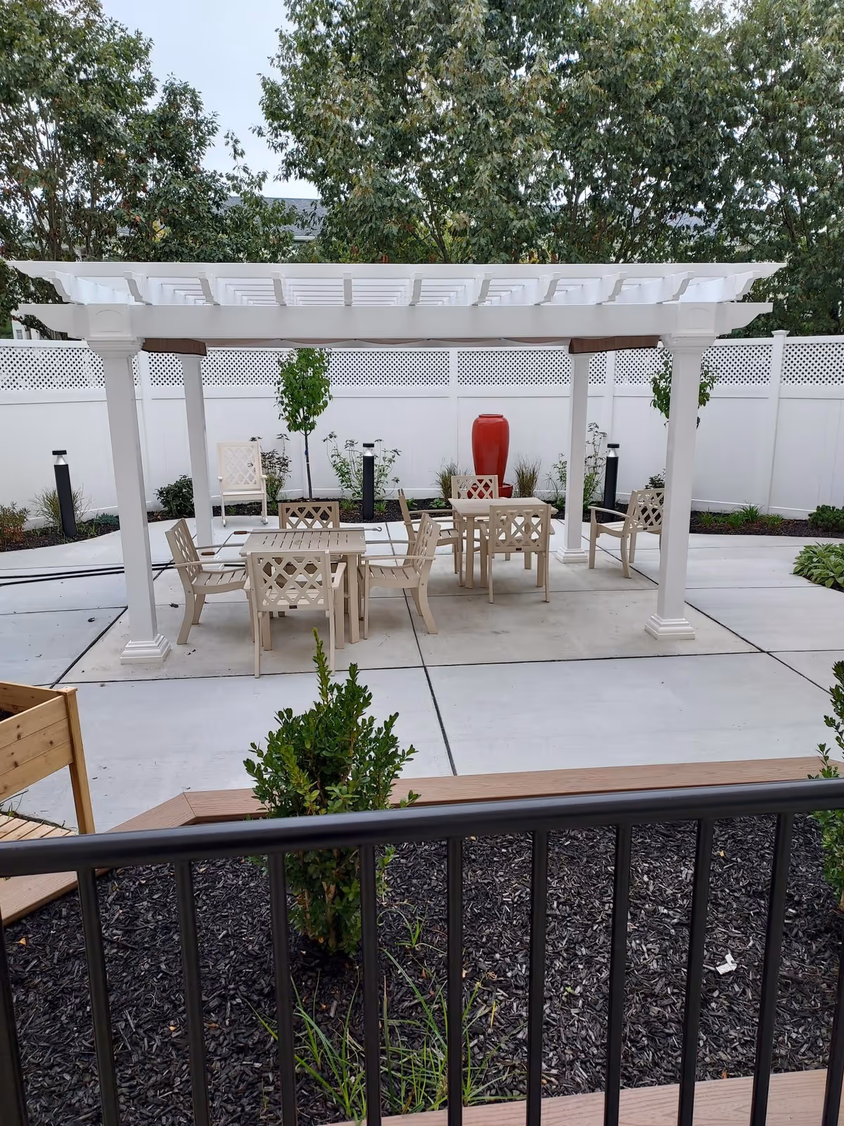 Outdoor patio with a white pergola over tables and chairs, landscaping, and a red decorative fountain by a white fence.