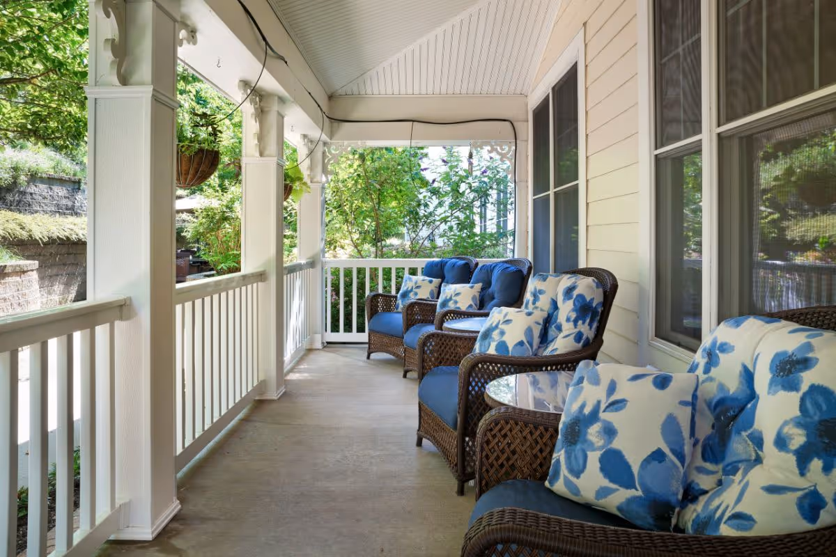 A covered porch with white railings and columns, featuring four wicker chairs with blue cushions and floral-patterned pillows. There is a small glass-top table between two of the chairs. Greenery and plants are visible beyond the porch.