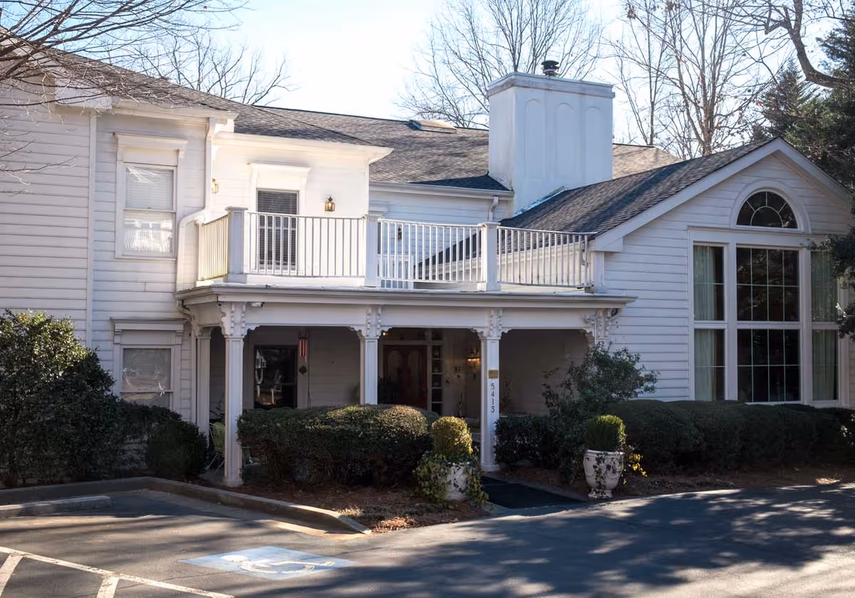 Exterior view of a two-story white building with a covered entrance, balcony, large windows, and surrounding bushes. There is a parking area with a handicapped parking space in front.