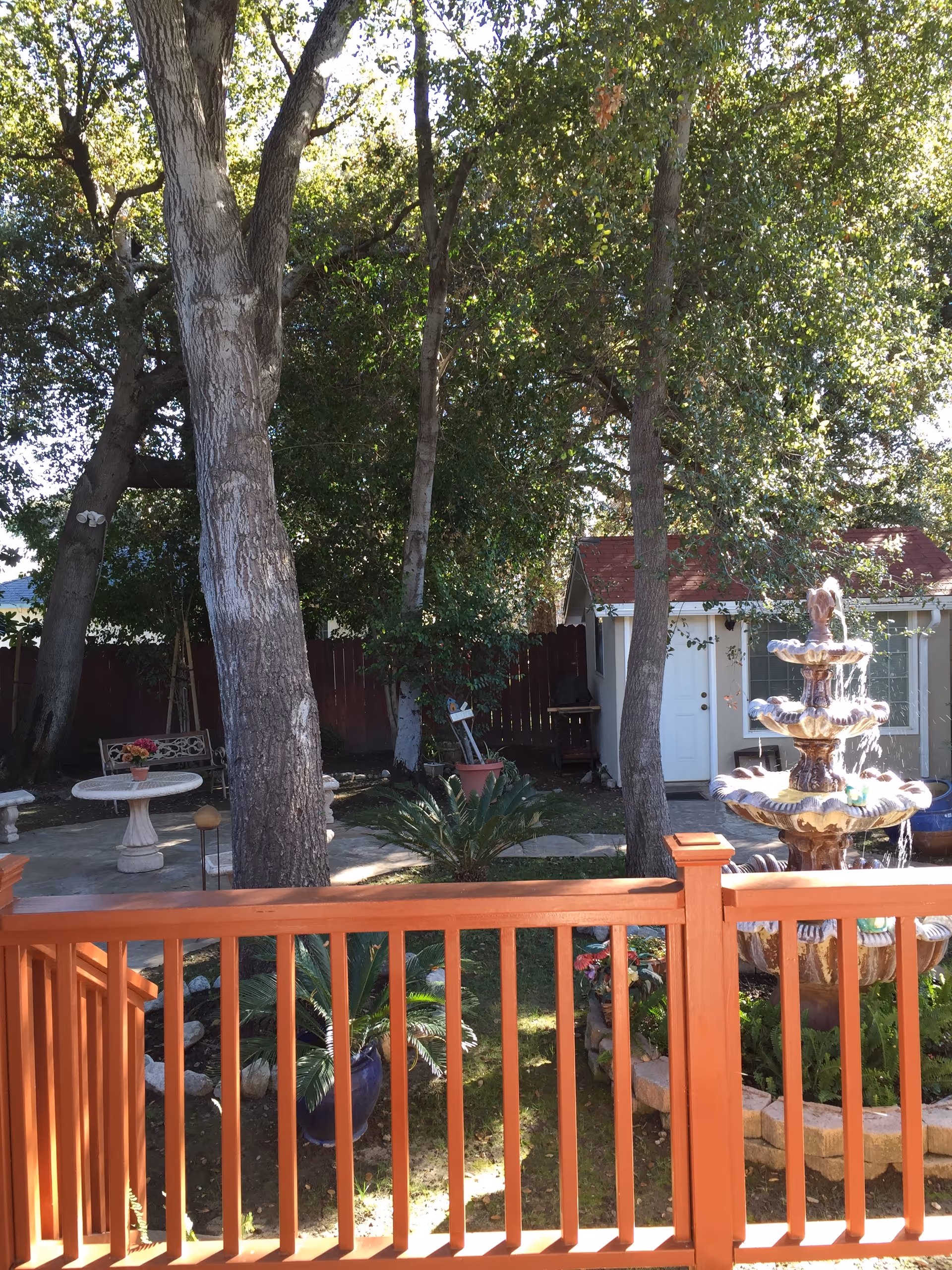 A peaceful outdoor garden area with large trees, a three-tiered water fountain, a small shed with a red roof, and a patio with a round table and bench. The foreground features a wooden railing painted in a reddish-brown color.