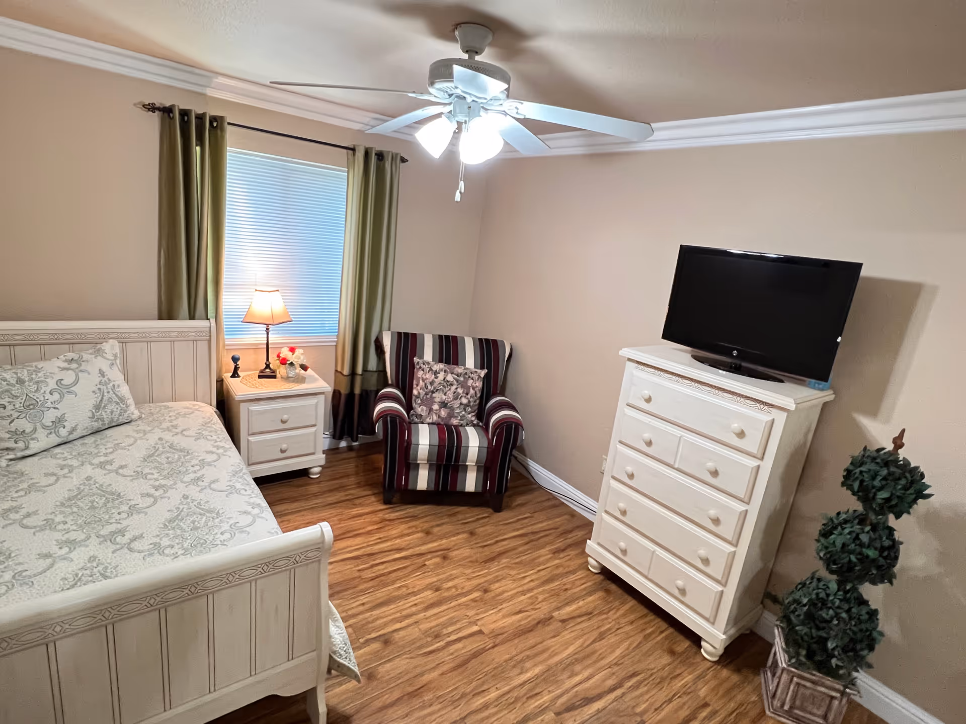 Neat bedroom with a white wooden bed and dresser, striped armchair, nightstand with a lamp, ceiling fan, and hardwood floor.
