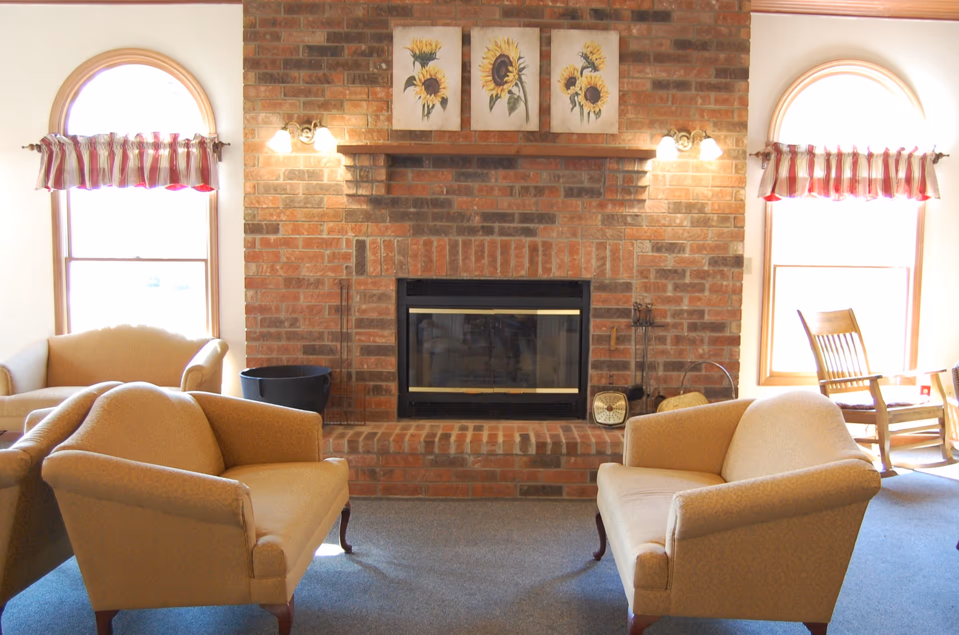 A cozy living room with a brick fireplace in the center, flanked by two arched windows with striped valance curtains. Two beige upholstered armchairs face each other in front of the fireplace, and a wooden rocking chair is positioned near the right window. Above the fireplace are three framed sunflower paintings, and wall-mounted lights illuminate the area.
