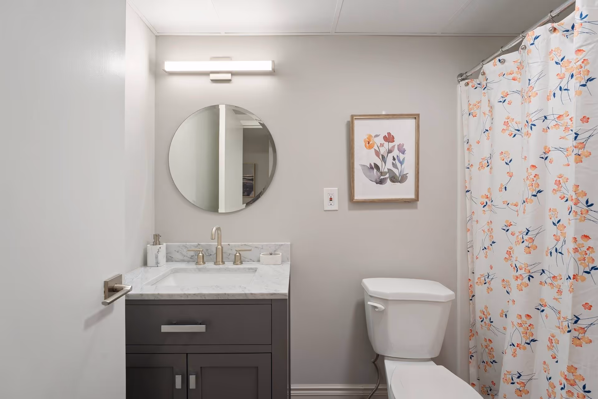 A clean, modern bathroom featuring a white toilet, a gray vanity with a marble countertop and built-in sink, a round mirror above the sink, a wall-mounted light fixture, a floral framed artwork on the wall, and a shower with a floral-patterned curtain.