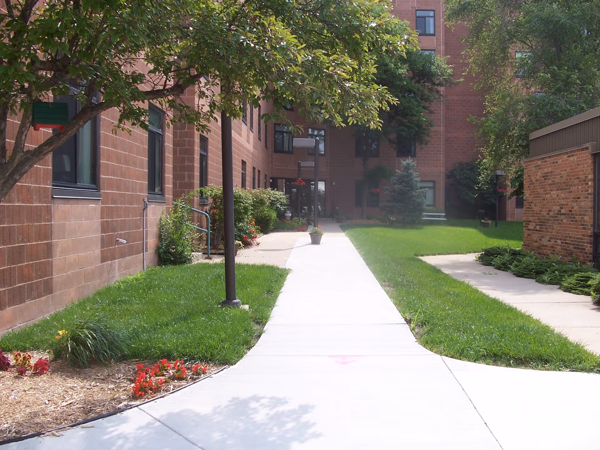 A paved walkway leading through a green courtyard area with grass, flower beds, and trees, surrounded by brick buildings. There are lamp posts along the path and a bench in the distance.