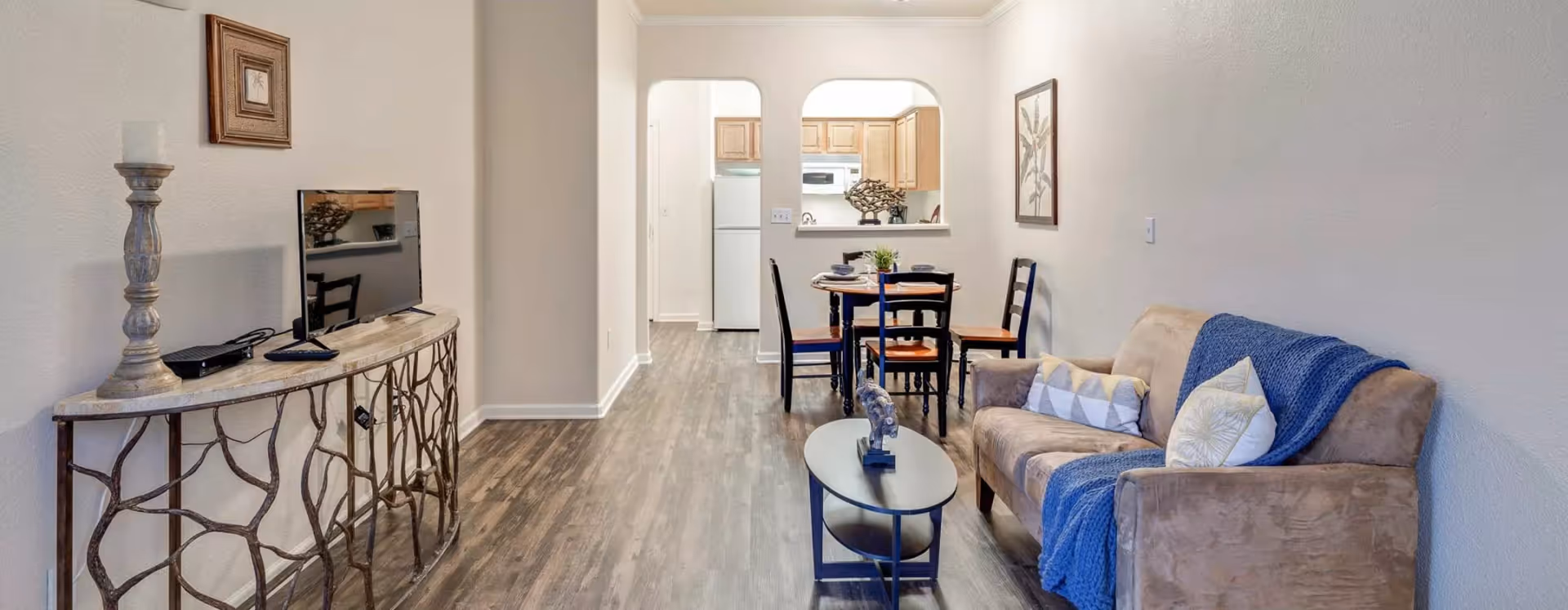 Open-plan living room with a sofa, coffee table, TV console and a small dining area leading to a kitchen.