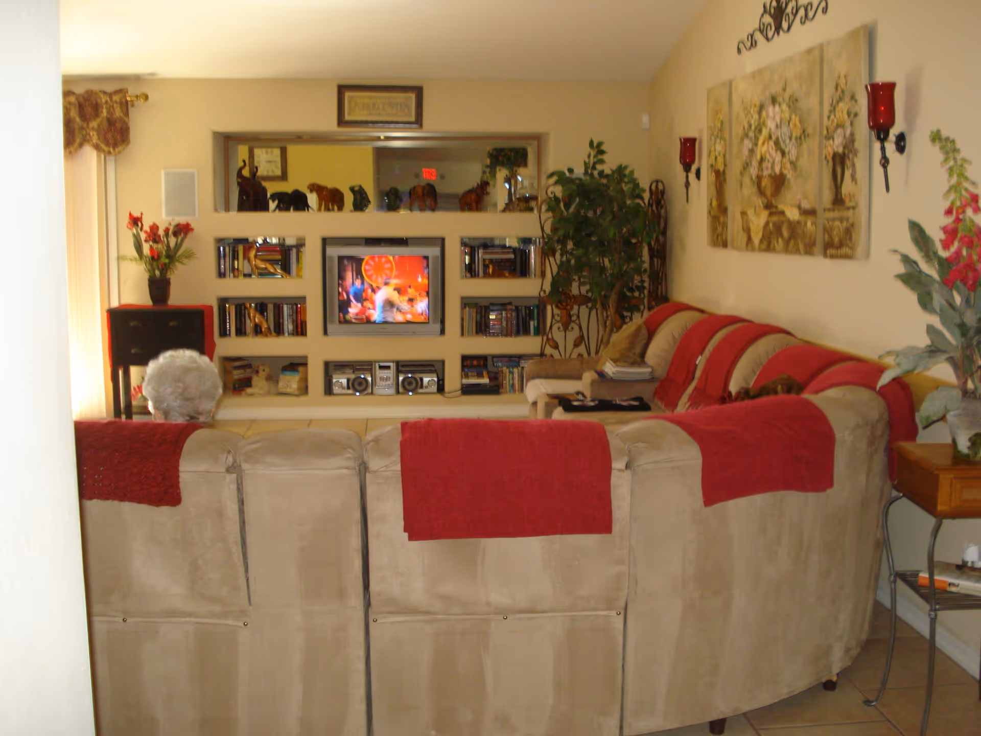 A cozy living room with a beige sectional sofa adorned with red throws. An elderly person with white hair is seated on the sofa facing a built-in entertainment center with a TV, books, and decorative elephant figurines. The room has warm yellow walls, floral paintings, wall sconces with red candles, and several potted plants.
