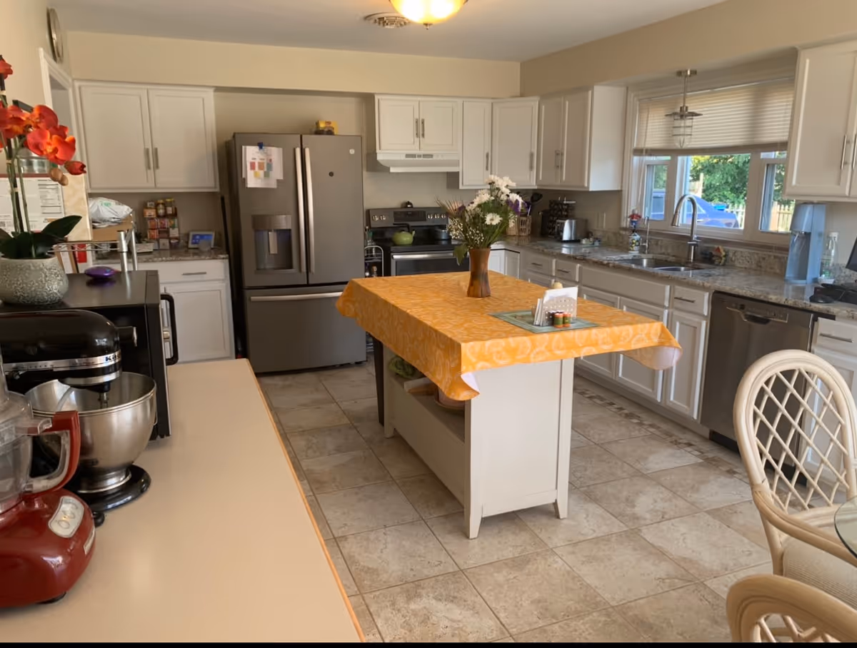 A bright and spacious kitchen with white cabinets, a stainless steel refrigerator, stove, and dishwasher. There is a kitchen island in the center covered with an orange tablecloth and a vase of flowers on top. Various kitchen appliances like a mixer and blender are on the countertops. A window above the sink lets in natural light, and a glass-top table with chairs is partially visible on the right.