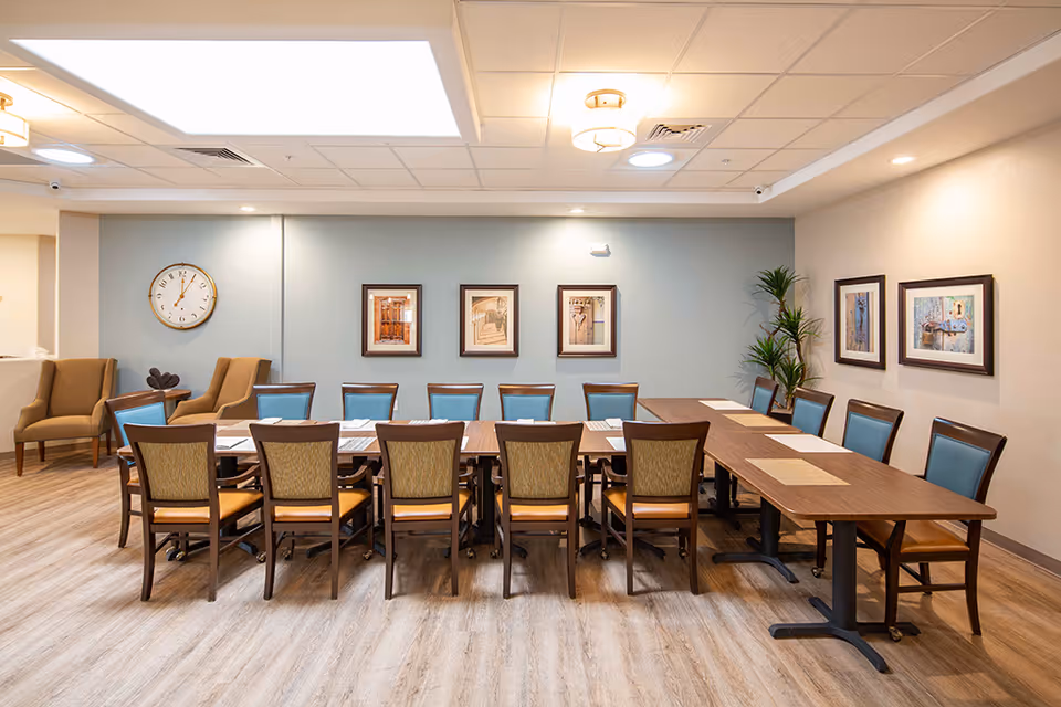Bright communal dining/activity room with a long wooden table surrounded by chairs, framed artwork on the walls, and a small seating area in the corner.