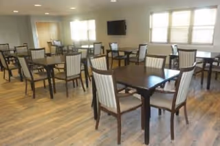 A spacious dining room with multiple square wooden tables and cushioned chairs arranged neatly. The room has large windows with blinds allowing natural light to fill the space. A flat-screen TV is mounted on the wall.