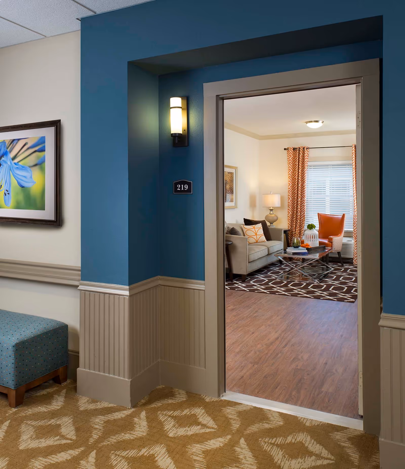 View through an open doorway into a cozy living room with a beige sofa, orange patterned pillows, an orange armchair, a glass coffee table on a dark patterned rug, and a window with orange curtains. The hallway outside the room has blue and beige walls, a wall-mounted light, a framed flower picture, and a blue upholstered bench.