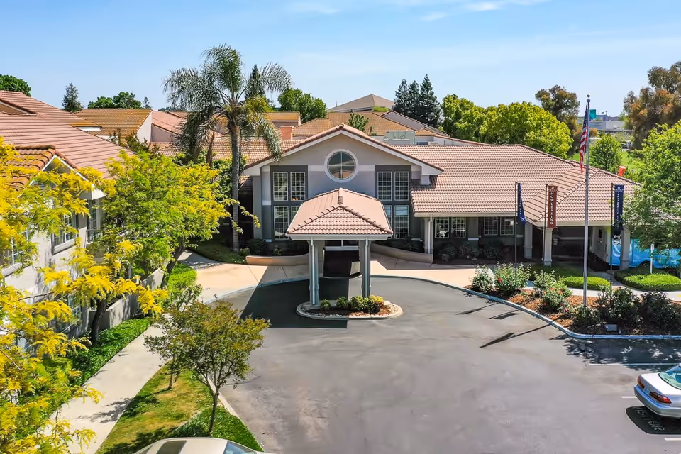 Front exterior view of Fresno Senior Living facility showing a building with a tiled roof, a covered entrance, surrounding greenery, and a circular driveway with parked cars.
