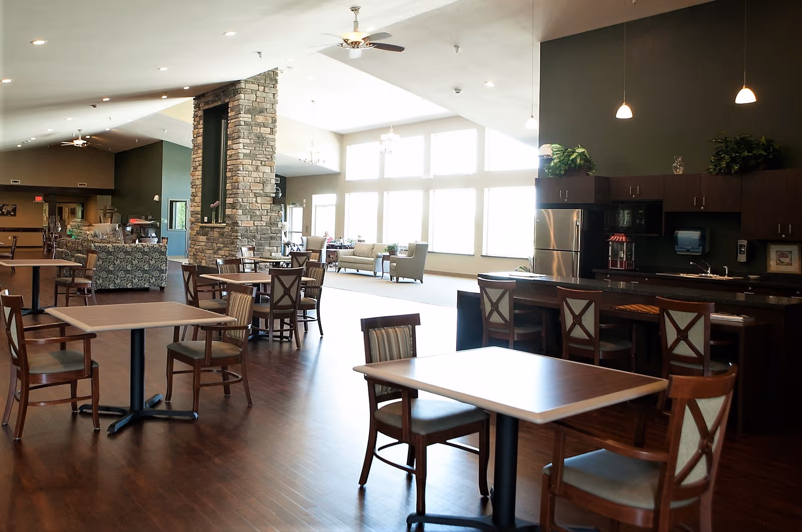 A spacious dining and common area in a senior living facility with wooden tables and chairs, a stone fireplace, large windows letting in natural light, and a kitchenette with a refrigerator and bar seating.