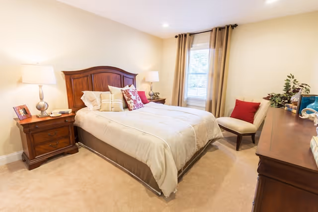 A cozy bedroom with a large wooden bed featuring beige bedding and multiple decorative pillows. There are two wooden nightstands with lamps on either side of the bed. A window with beige curtains lets in natural light. A cushioned chair with a red pillow is placed near a wooden dresser adorned with decorative items and plants.