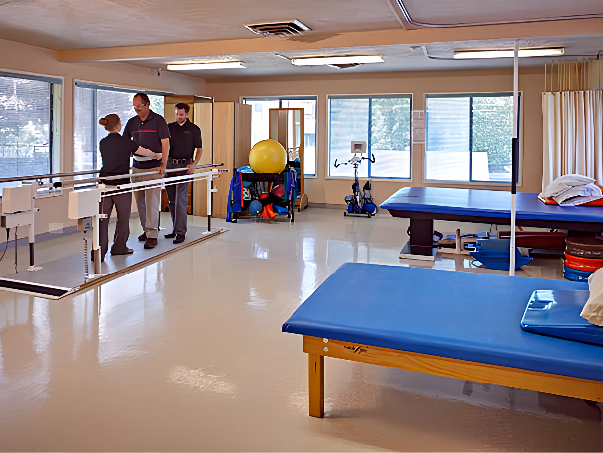 A physical therapy room with two therapists assisting a man walking between parallel bars. The room has large windows, exercise equipment including a stationary bike, therapy tables with blue padding, and various therapy balls and supplies.