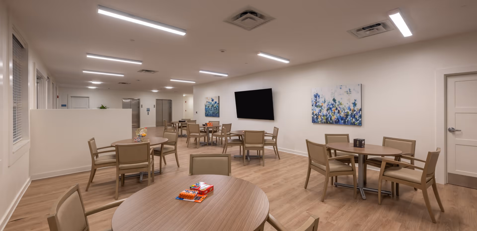 Bright communal dining/activity room with round tables and chairs, a wall-mounted TV, and artwork on the walls.