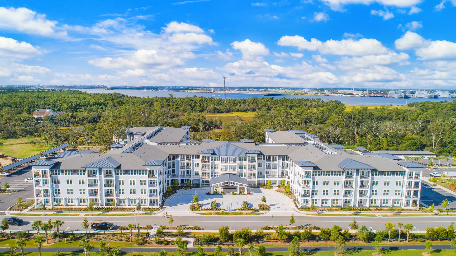 Aerial view of a large, modern senior living facility building with multiple floors and a central entrance, surrounded by parking lots and greenery. In the background, there is a dense forest area and a body of water under a partly cloudy blue sky.