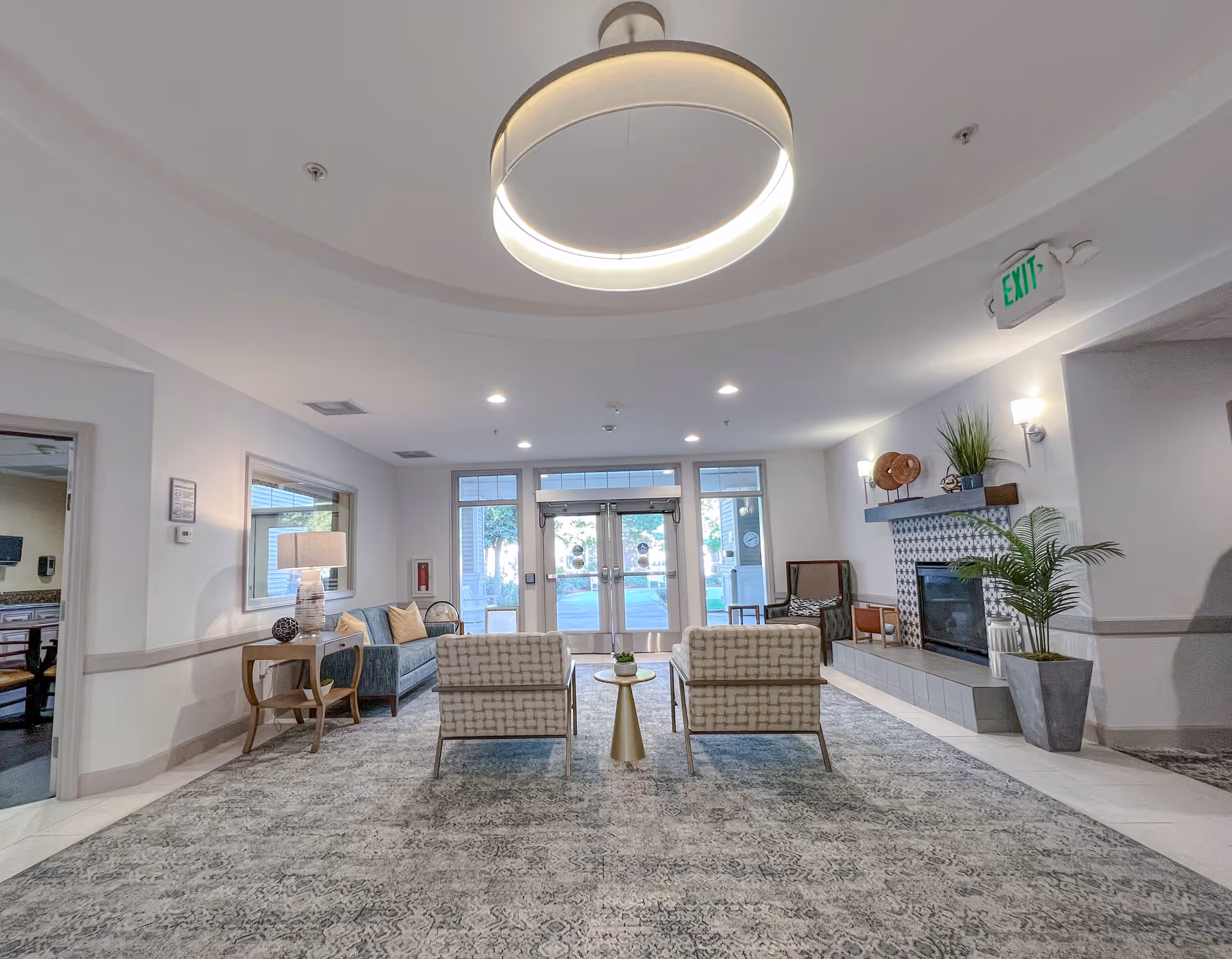A bright and spacious living room area with two patterned armchairs facing a small round table, a blue sofa with cushions, a side table with a lamp, a fireplace with decorative items on the mantel, potted plants, and large glass doors leading outside. The ceiling has a modern circular light fixture and recessed lighting.