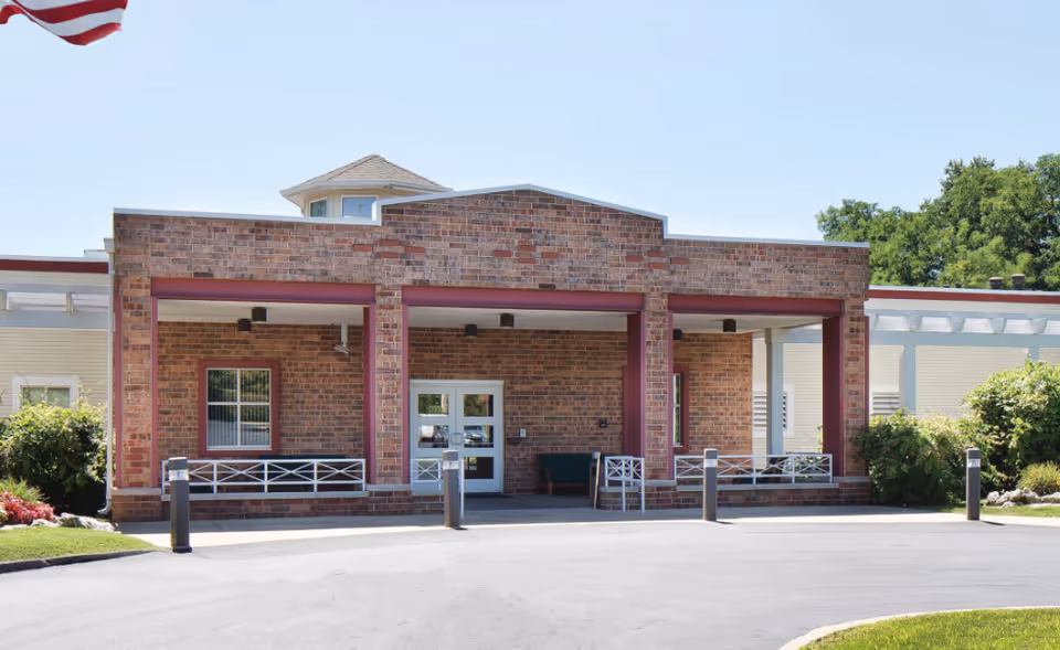 Front exterior view of a single-story brick building with a covered entrance supported by red columns. There are white benches on the porch area and a glass door entrance. An American flag is partially visible in the upper left corner, and there are green bushes and trees surrounding the building.