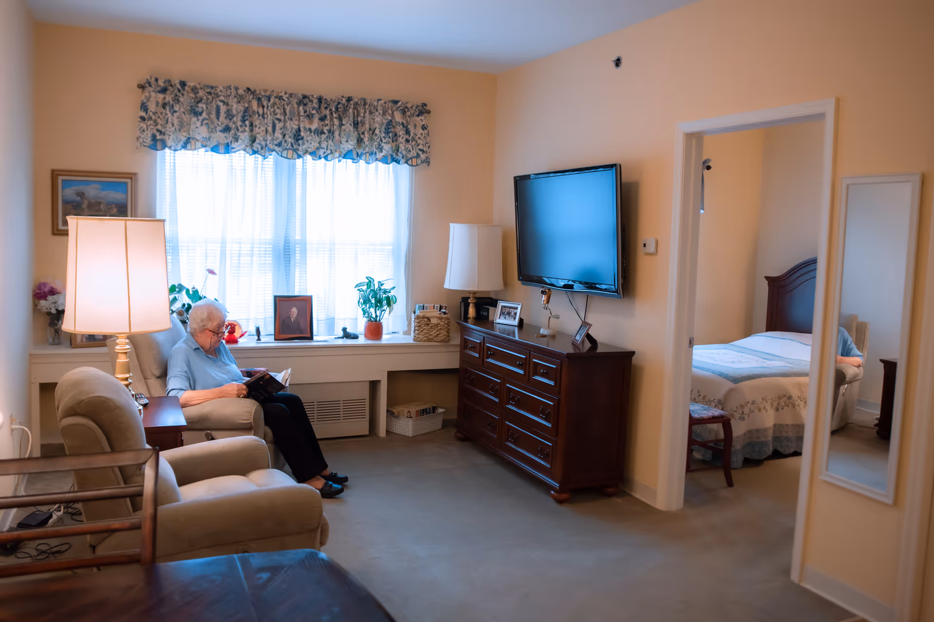 An elderly woman sits reading in a sunlit living room with a TV on a dresser and a bedroom visible through an open doorway.