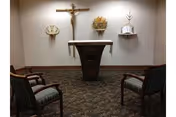 A small chapel room with a crucifix mounted on the wall above a wooden altar. There are three religious symbols displayed on the wall behind the altar. Two wooden chairs with striped cushions are positioned facing the altar on a patterned carpeted floor.