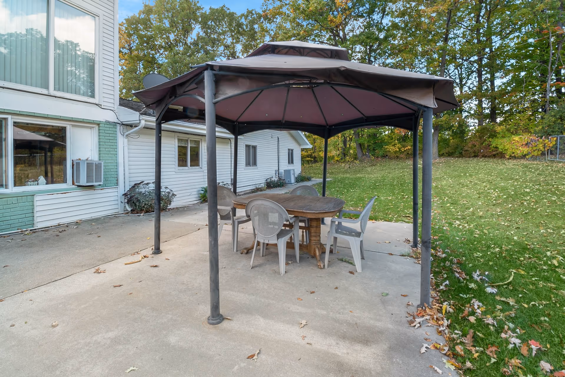 Outdoor patio area with a gazebo covering a wooden table and four plastic chairs. The patio is adjacent to a white building with green brick accents and surrounded by grass and trees with autumn foliage.