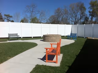 Outdoor seating area with a circular brick fire pit in the center, surrounded by a concrete walkway and three benches painted in orange, blue, and black. The area is enclosed by a tall white fence with trees visible in the background under a clear blue sky.