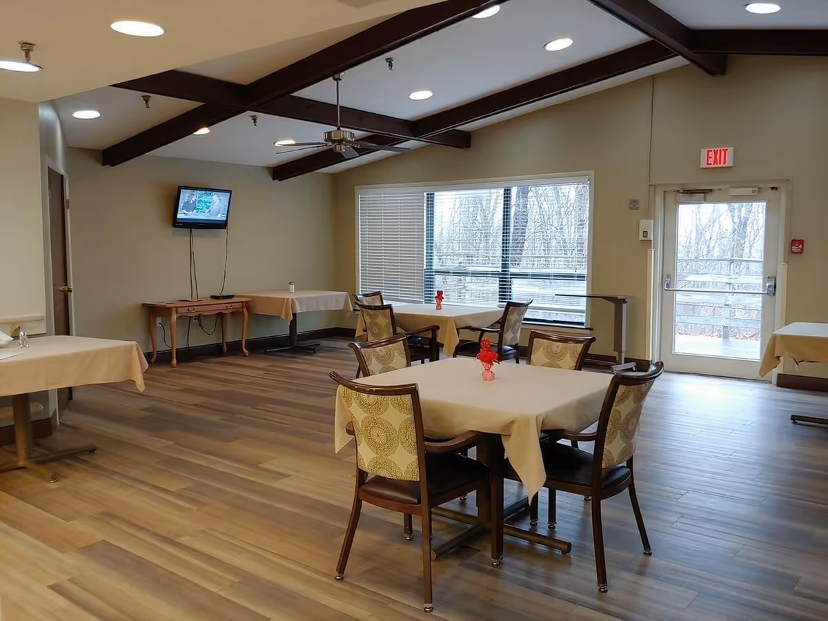 A dining room with several tables covered with beige tablecloths and chairs with patterned upholstery. The room has wooden flooring, a ceiling fan, exposed dark wooden beams, and a wall-mounted TV. Large windows and a glass door let in natural light and show a view of trees outside.