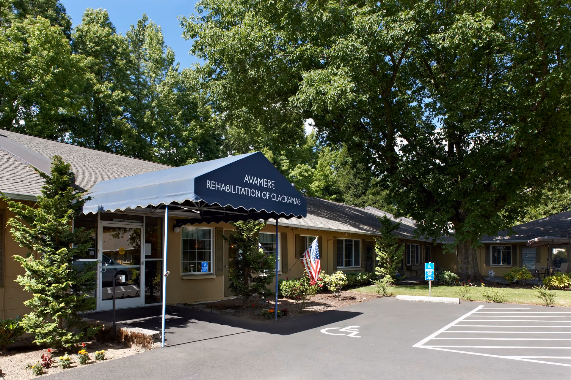 Front entrance of Avamere Rehabilitation of Clackamas featuring a blue awning over the doorway, landscaped beds, an American flag, and a marked handicap parking space.