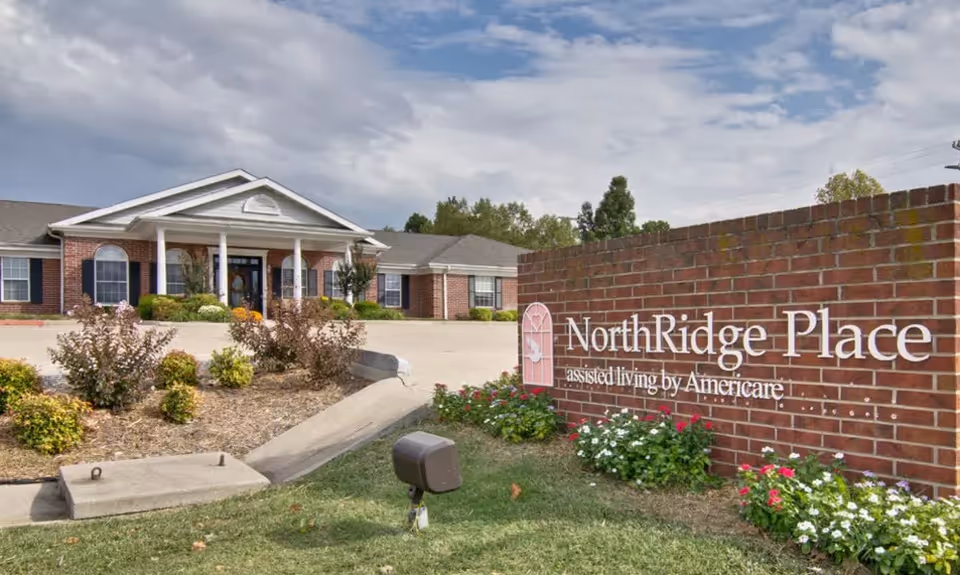 Exterior view of NorthRidge Place assisted living facility showing a brick sign with the facility name and a large building with white columns and a landscaped garden under a partly cloudy sky.