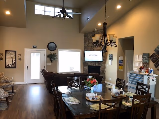 Community dining and living area with a large wooden dining table and chairs in the foreground, sofas and a TV above a stone fireplace in the background, and high ceilings with a fan and chandelier.