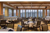Spacious dining room with multiple round tables covered with white tablecloths and set with napkins and glassware. The room features large windows along one wall, wooden ceiling beams, and a warm, inviting atmosphere with cushioned chairs arranged around the tables.