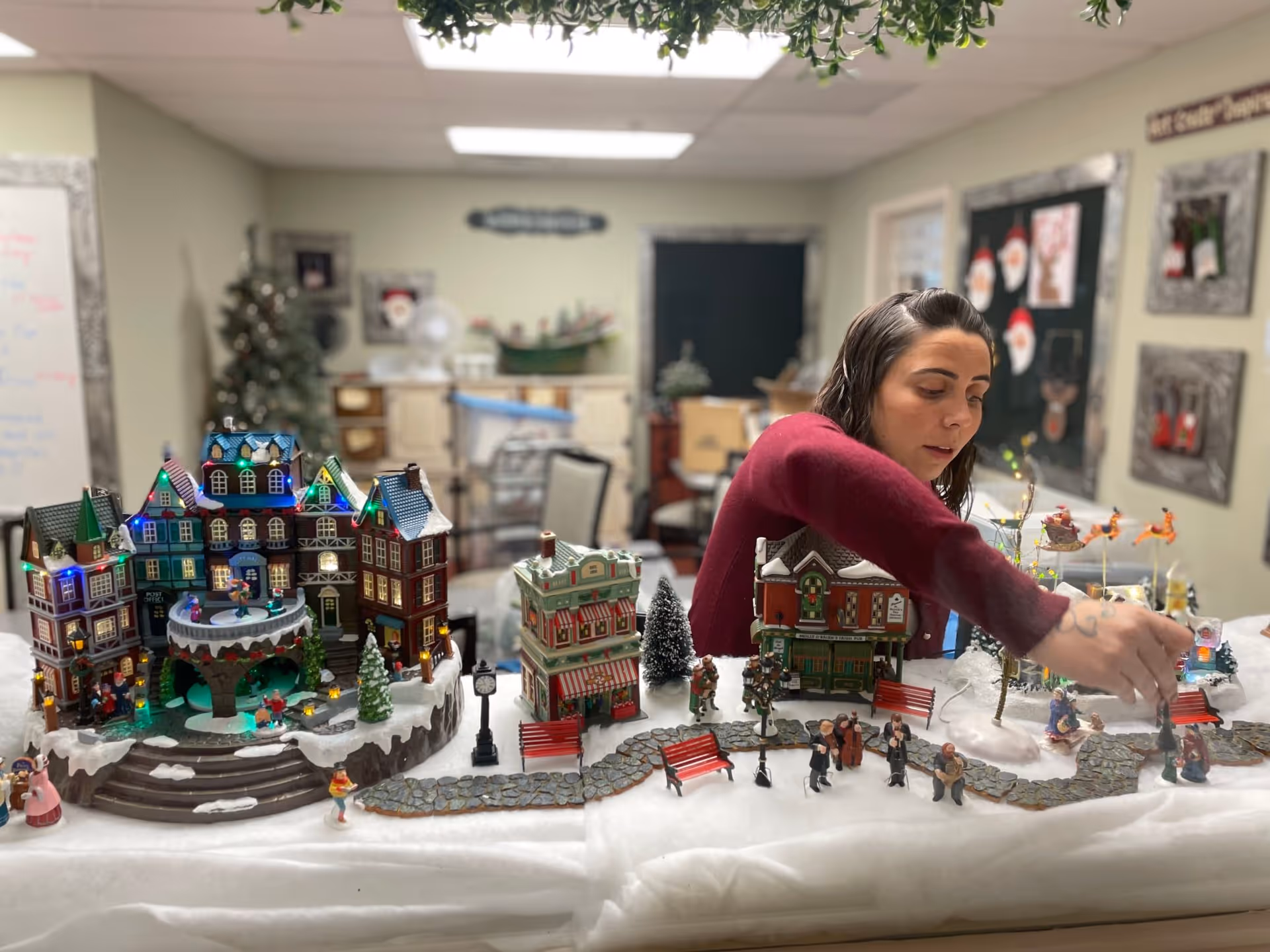 A woman in a red sweater arranges a miniature Christmas village display on a table covered with white fabric. The village features small buildings with colorful lights, figurines of people, benches, trees, and a pathway. The background shows a room with chairs, a Christmas tree, and holiday decorations on the walls.