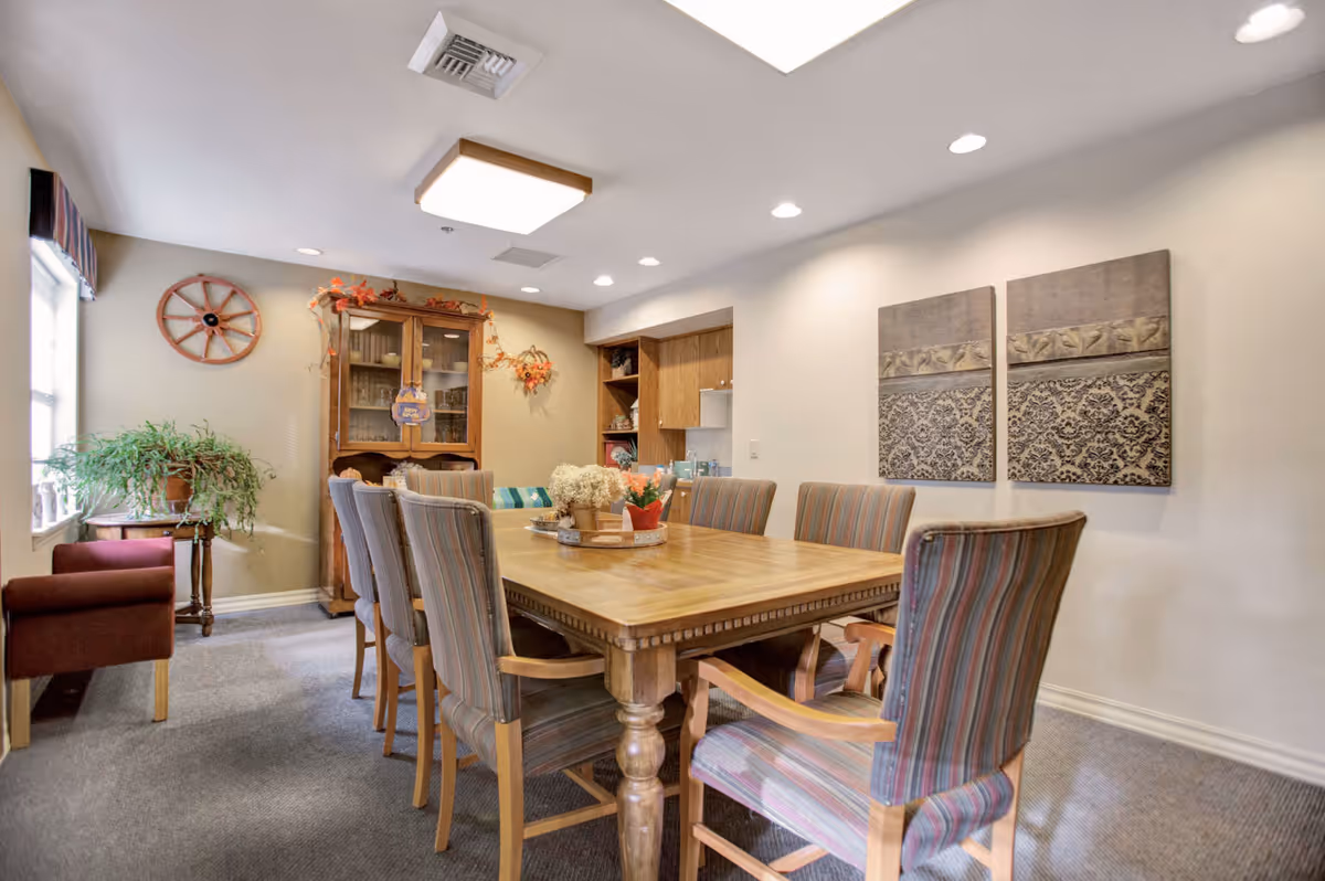 A well-lit dining room with a wooden table surrounded by eight striped upholstered chairs. The room features a wooden cabinet with glass doors, decorative wall art, a plant on a small table near a window, and a comfortable armchair. The walls are painted light beige, and the ceiling has recessed lighting and a central square light fixture.