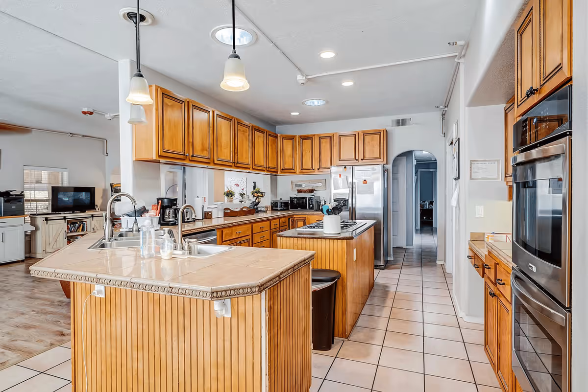 A spacious kitchen with wooden cabinets and tiled countertops. The kitchen features a double sink, a central island with a cooktop, stainless steel refrigerator, built-in double oven, and various small appliances like a coffee maker and microwave. The floor is tiled, and there are pendant lights hanging from the ceiling. An archway leads to another room in the background.