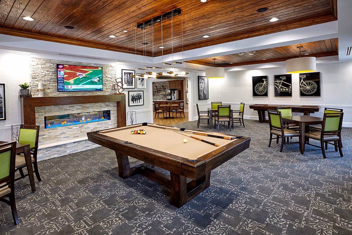 A spacious recreational room featuring a pool table with cues and balls set up, several tables with green cushioned chairs, a modern electric fireplace with a mounted flat-screen TV above it showing a sports game, and decorative framed artwork on the walls. The room has a patterned carpet, wooden ceiling accents, and pendant lighting.