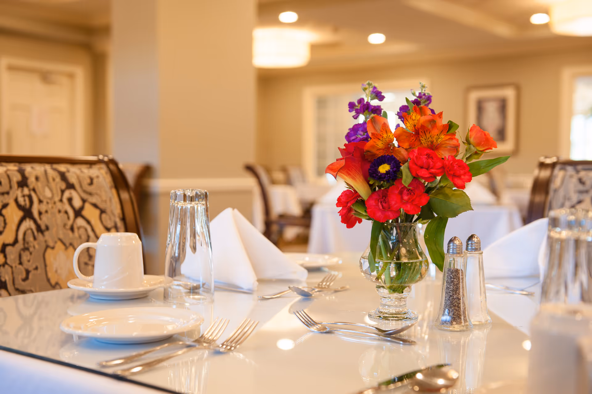 A dining table set with white plates, silverware, a white cup on a saucer, an upside-down glass, folded white napkins, and a vase of colorful flowers including red, orange, and purple blooms. The background shows a softly lit dining room with patterned chairs and framed artwork on the wall.