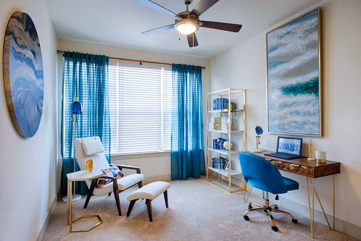 A bright room with a large window covered by blue curtains and white blinds. The room features a white armchair with a matching footrest, a small side table with a glass of orange juice, a blue swivel chair at a wooden desk with a laptop, and a gold and white shelving unit holding decorative items and books. The walls are adorned with two abstract paintings, and a ceiling fan with a light is mounted above.